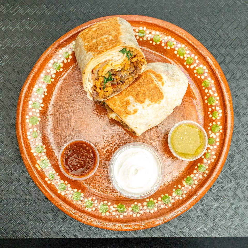 Two burritos, one cut open, revealing meat and rice, served with three dipping sauces on a decorative plate.