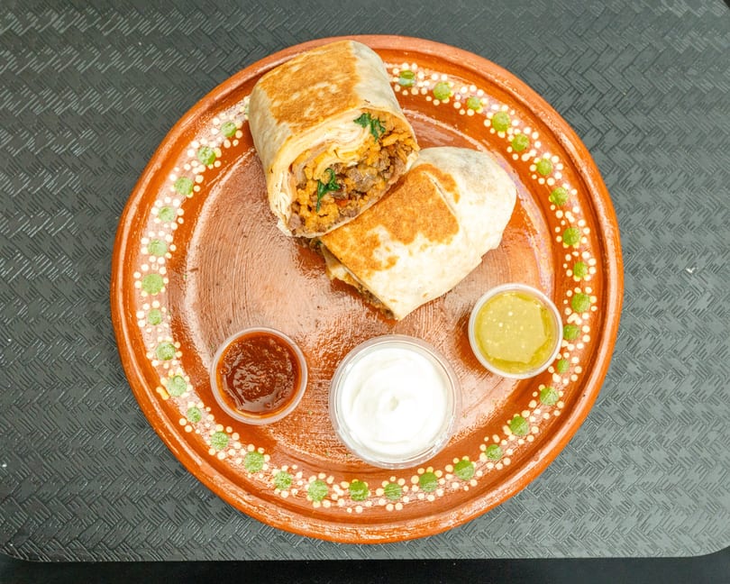 Two burritos, one cut open, revealing meat and rice, served with three dipping sauces on a decorative plate.