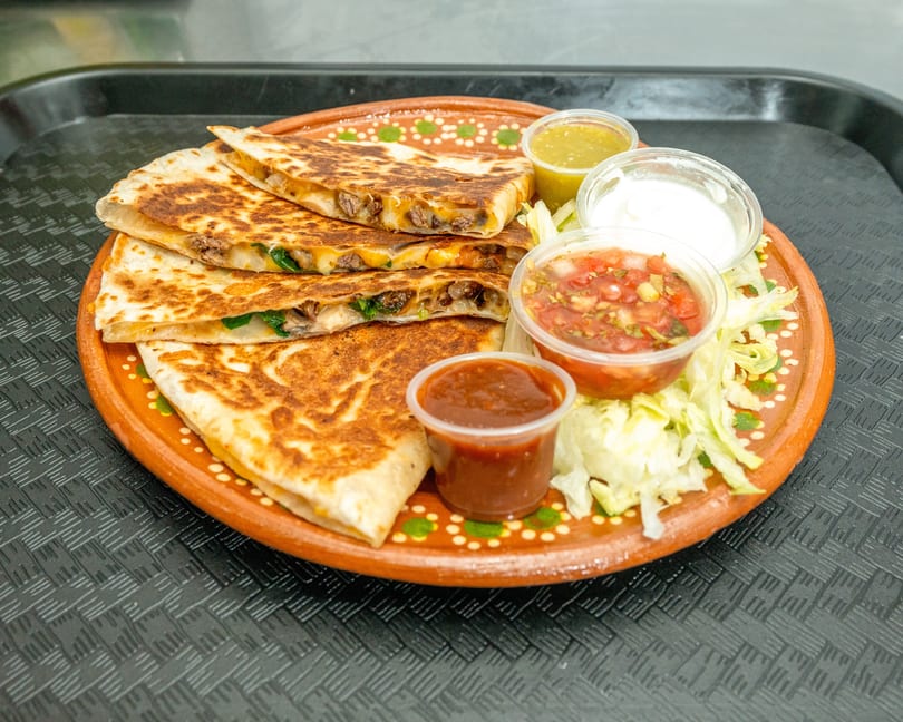 Four quesadilla slices with meat and cheese, served with salsa, sour cream, and shredded lettuce.