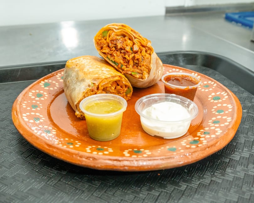 Two burritos, one cut in half, on a decorative orange plate with three dipping sauces.